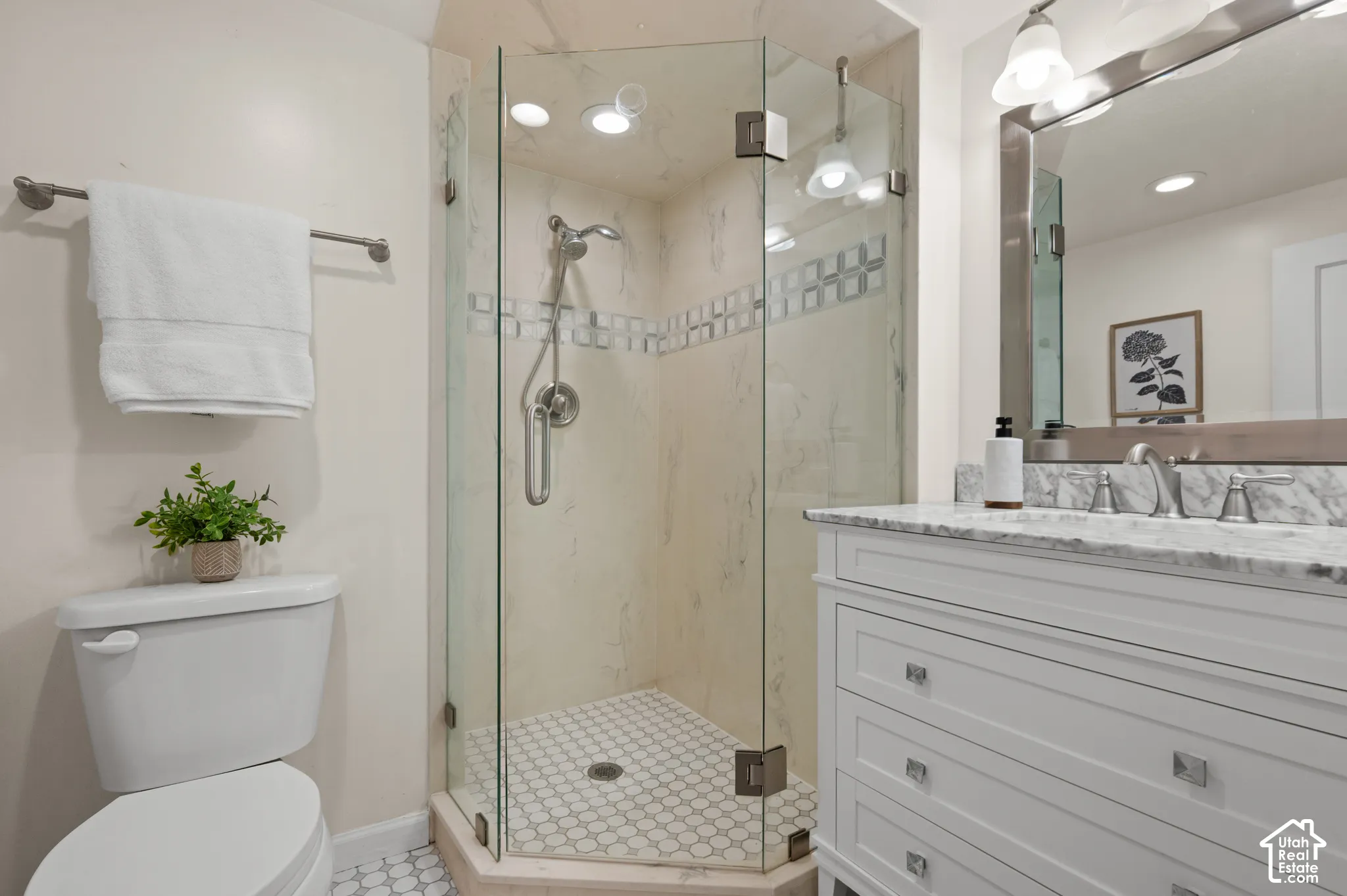 Corner shower enclosure with glass walls and decorative tile inlay, white vanity with stone-finish countertop, undermount sink, and polished nickel hardware