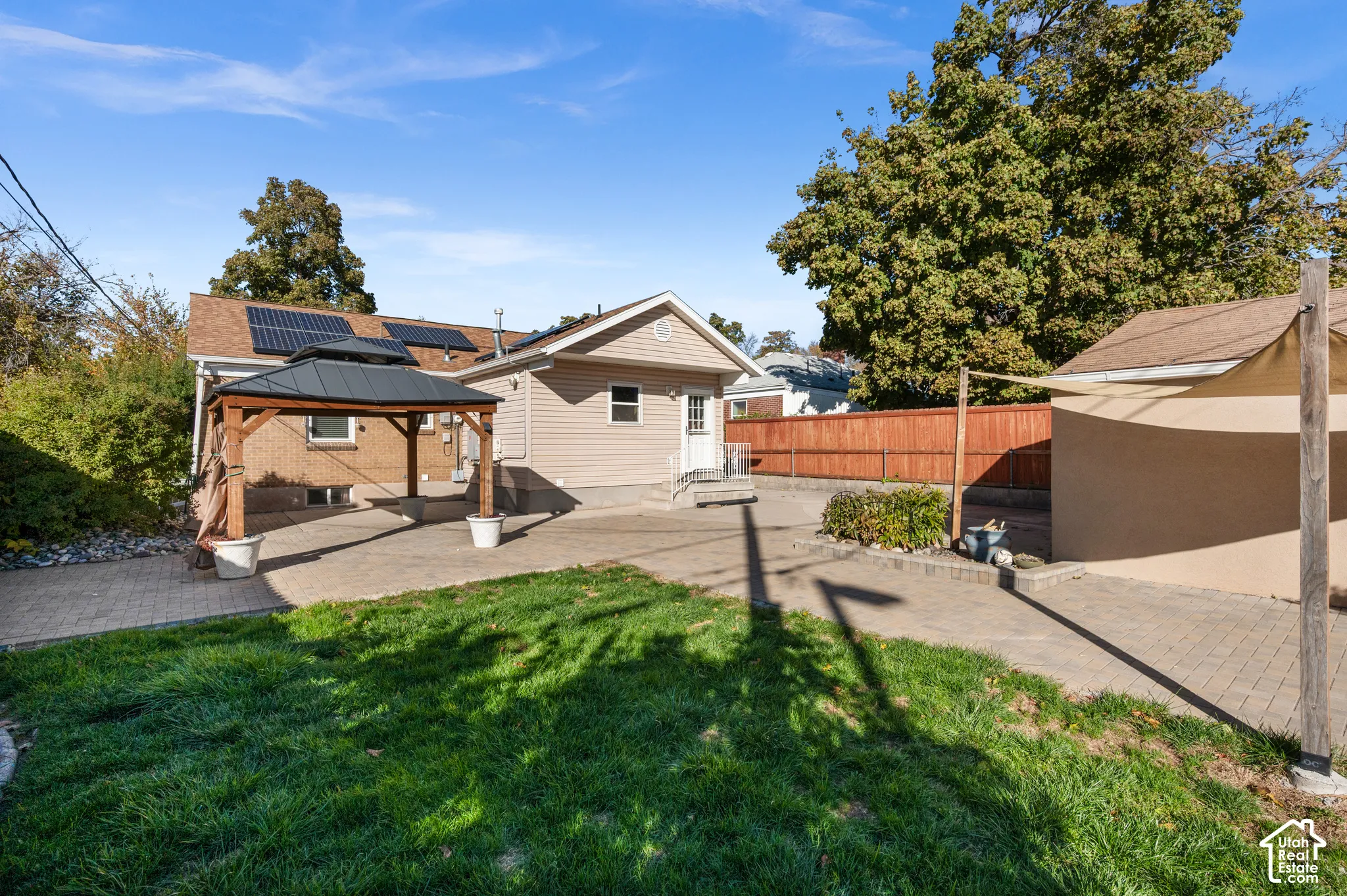 Exterior space featuring a paved stone patio, permanent metal-roof gazebo, solar panel array, wood-fencing perimeter, and mature landscape trees