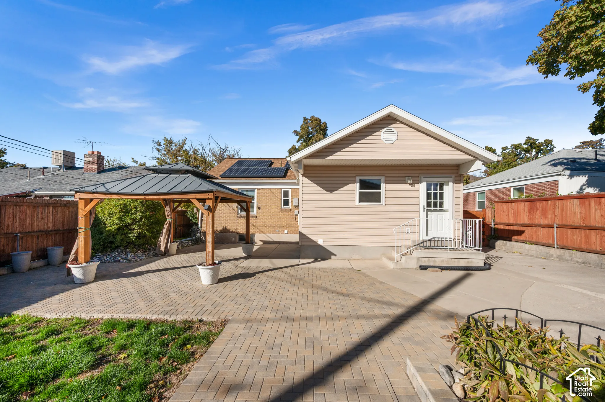 Rear exterior featuring light-toned vinyl siding, a paved brick patio, a fixed wooden gazebo, solar panel array, and full-perimeter wood fencing
