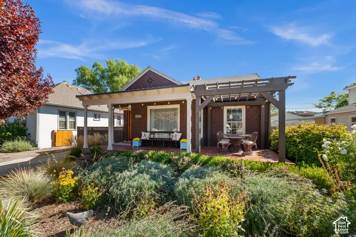 Brick facade residence featuring a covered porch, an open pergola, a brick patio, and mature landscaping