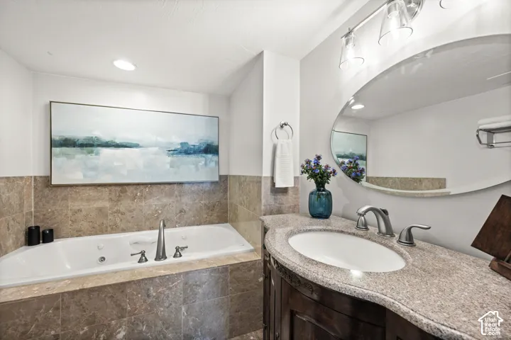 Soaking tub with tile surround, built-in vanity with an undermount sink, stone countertop, and an oval mirror