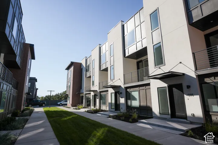 Modern multi-story exterior featuring a mix of stucco and brick siding, metal awning accents, balcony railings with horizontal cabling, and floor-to-ceiling windows