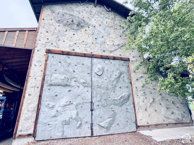 Exterior rock climbing wall with integrated holds, featuring large double doors and a wood-framed canopy - backside of barn building.
