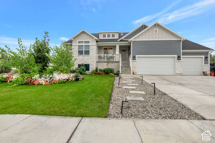 Stone and siding exterior with a large front lawn, landscaped garden beds, a front porch, and a multi-car garage