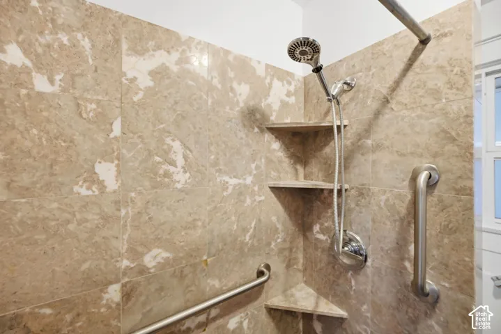 Shower stall with light-toned, variegated tile, built-in corner shelving, a handheld showerhead, and grab bars