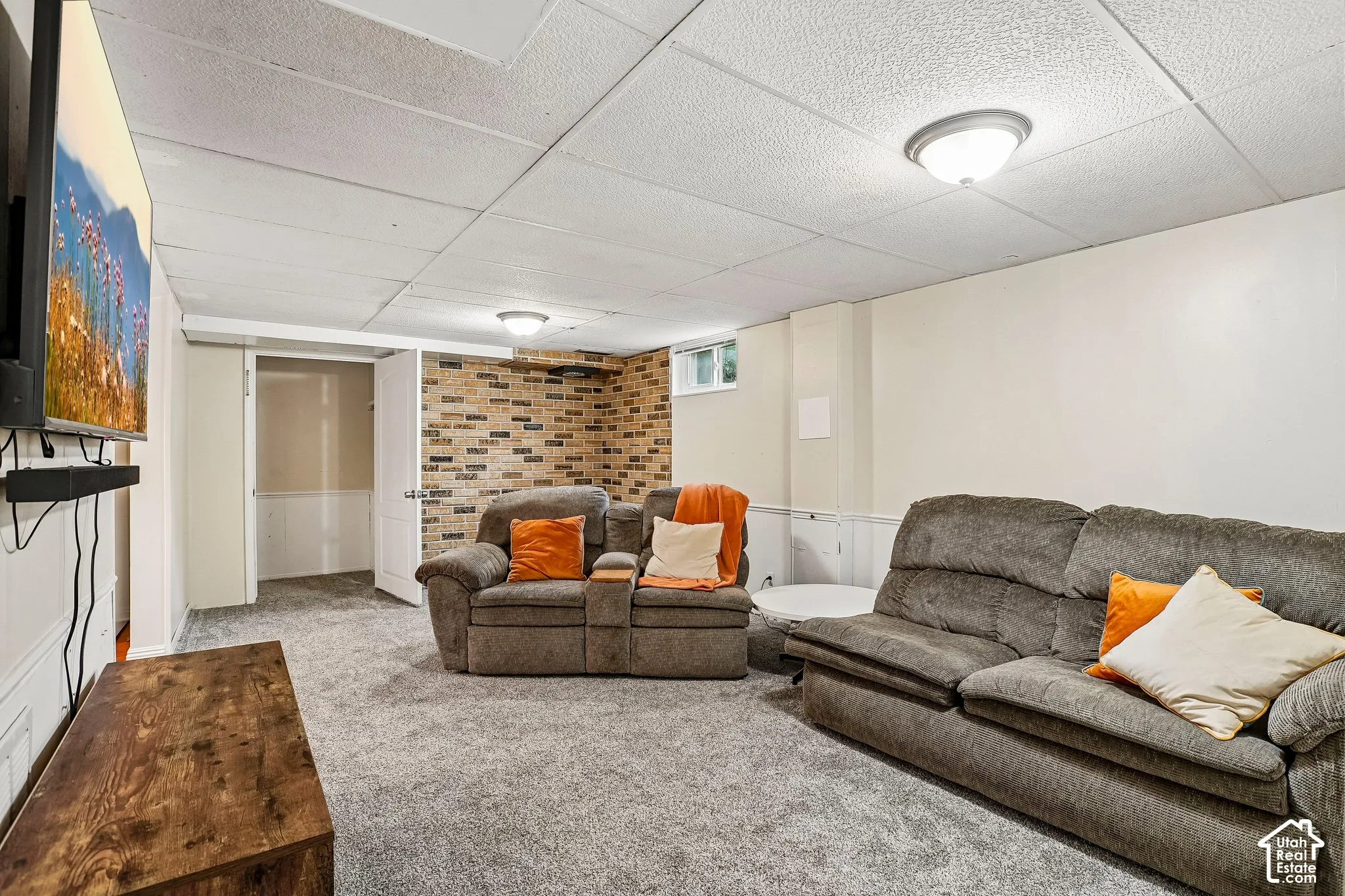 Recreation room featuring carpet flooring, drop ceiling with flush-mount lighting, and an accent wall with brick-finish paneling