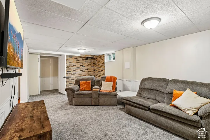 Recreation room featuring carpet flooring, drop ceiling with flush-mount lighting, and an accent wall with brick-finish paneling