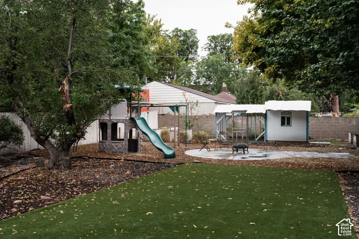 Expansive backyard featuring artificial turf, mature trees, a built-in fire pit area with a concrete patio, and a dedicated play structure
