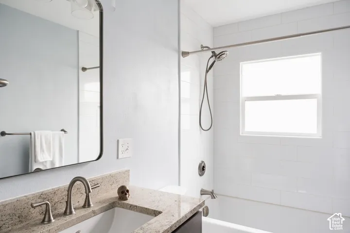 Bathroom featuring a rectangular undermount sink, stone-finish countertop, brushed nickel faucet, framed mirror, and a tiled shower with window