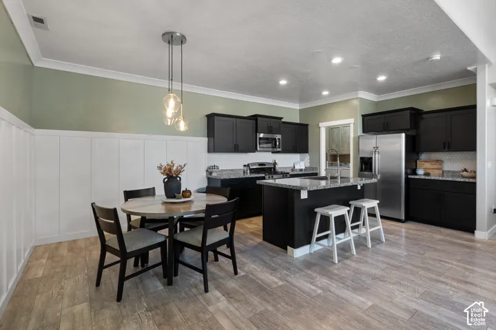 Open-concept kitchen and dining area featuring black cabinetry with stainless steel appliances, a center island with seating, wood-finish flooring throughout, sage green accent walls, white shiplap wainscoting, and recessed ceiling lighting with pendant f