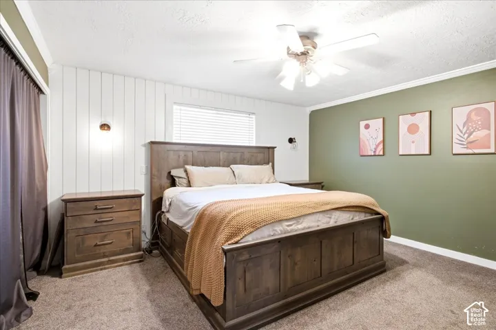 Textured carpet flooring, white shiplap accent wall, mounted wall sconces, ceiling fan with light fixture, and window with blinds