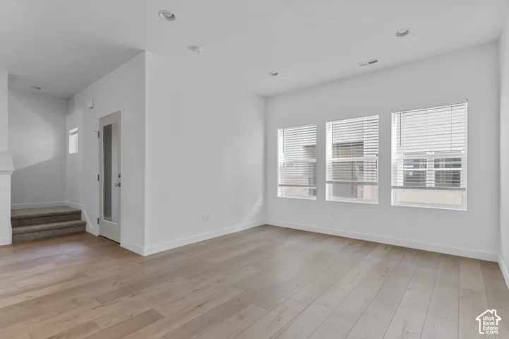 Light-toned wood-finish flooring, recessed ceiling lighting, three windows with blinds, white walls, and a staircase with carpeted treads