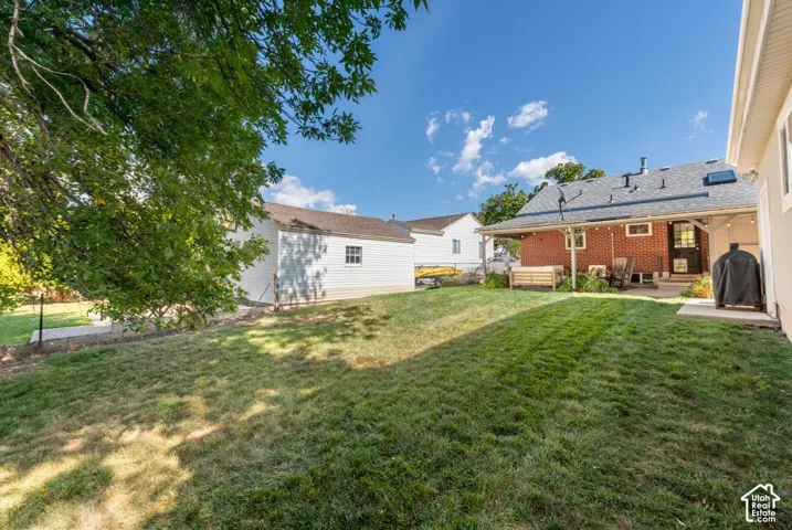 Expansive backyard featuring a large green space, a brick exterior house with a covered patio, a detached white outbuilding, and mature trees