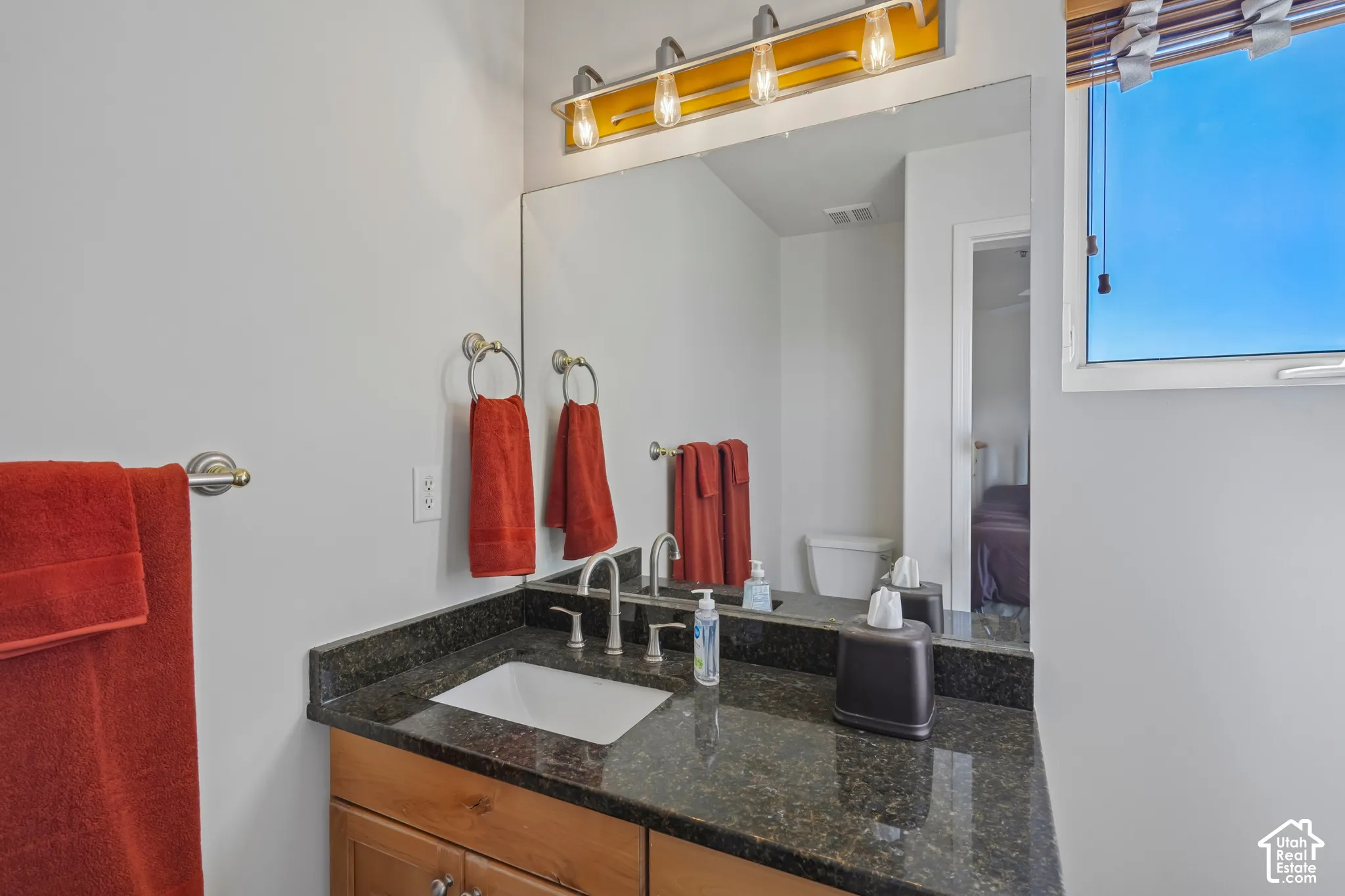 Dark-toned countertop with an integrated sink and chrome faucet, wood-finish vanity with drawer pulls, and a large mirror with overhead lighting