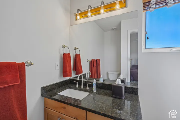 Dark-toned countertop with an integrated sink and chrome faucet, wood-finish vanity with drawer pulls, and a large mirror with overhead lighting