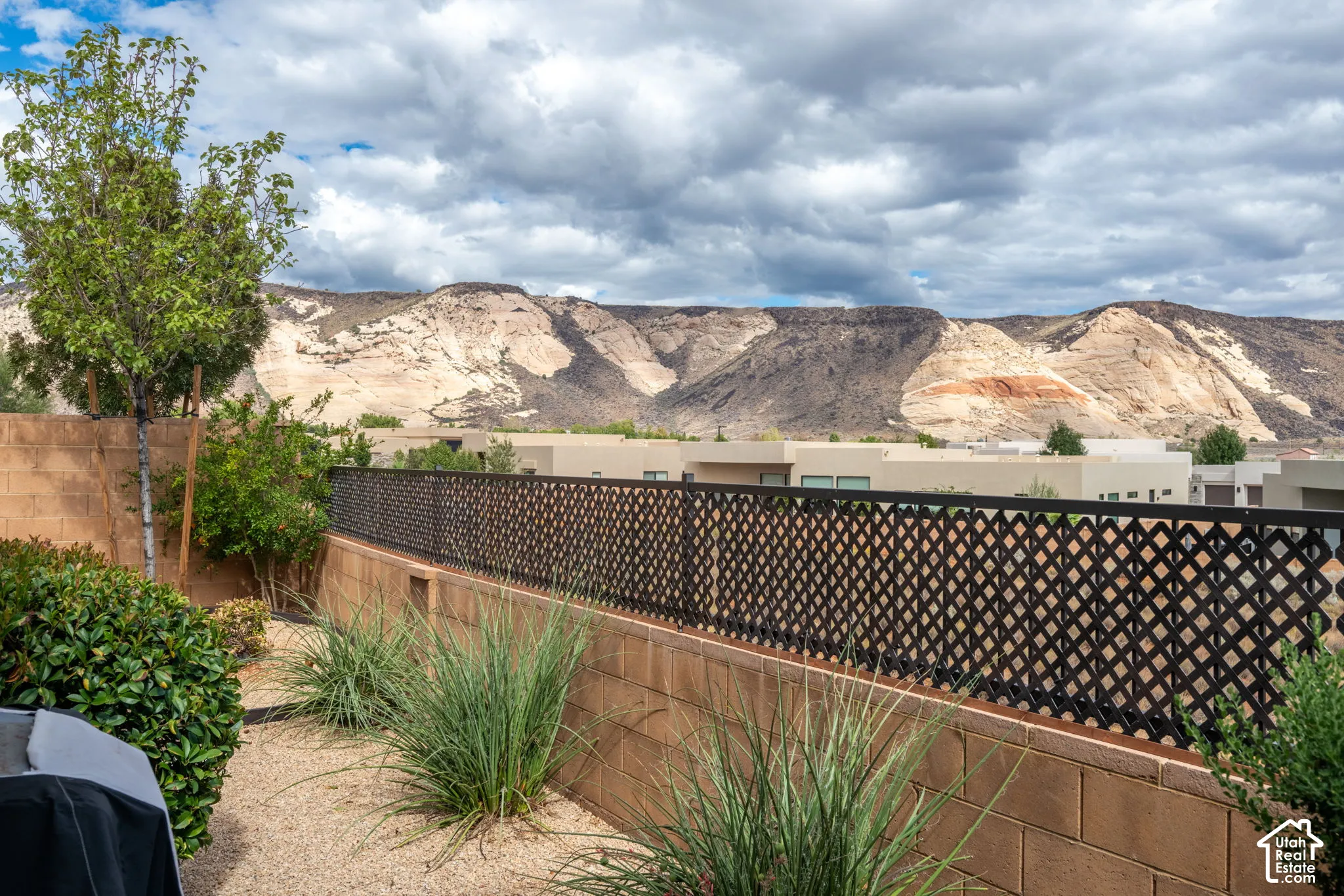 Lattice-patterned metal fencing with a concrete block retaining wall, desert landscaping featuring gravel and various plants, and distant mountainous terrain