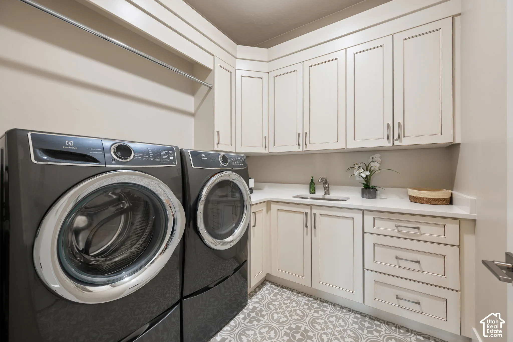 Laundry area featuring extensive white cabinetry, a built-in sink with a gooseneck faucet, light-toned countertops, and patterned floor tiles