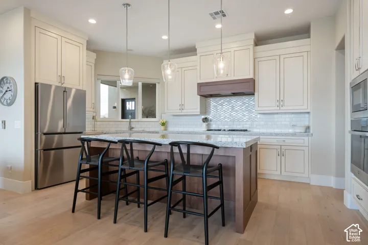 Kitchen featuring light wood-finish flooring, white cabinetry, a wood island base with a light-colored countertop, stainless steel appliances, and a tiled backsplash