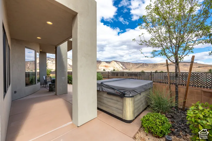 Covered patio with polished concrete flooring opening to a resort-style backyard featuring a built-in hot tub with surround decking, drought-resistant landscaping with ornamental grasses and shrubs, wooden privacy fencing, and panoramic mountain and valle