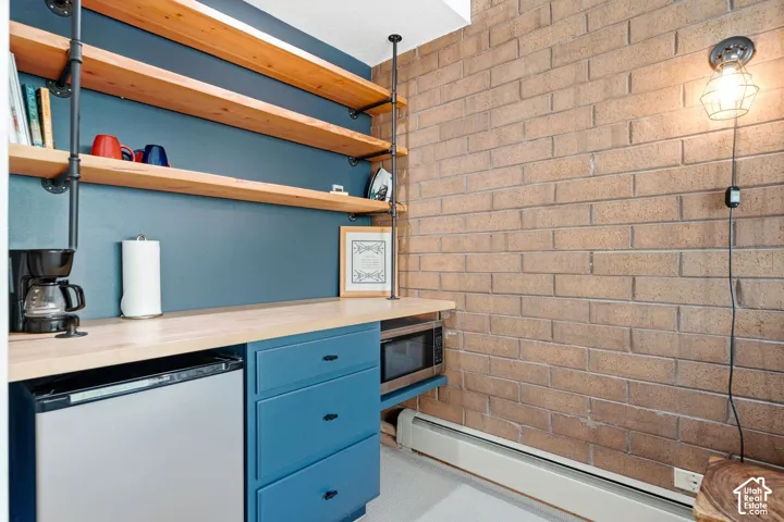 Built-in shelving with wood planks, painted base cabinets with drawers, wood-finish countertop, exposed brick wall, and wall-mounted light fixture