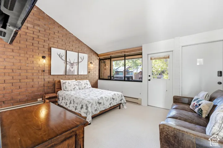 Exposed brick accent wall, sloped ceiling, wood-frame windows, light-tone carpet, and a built-in wall heater