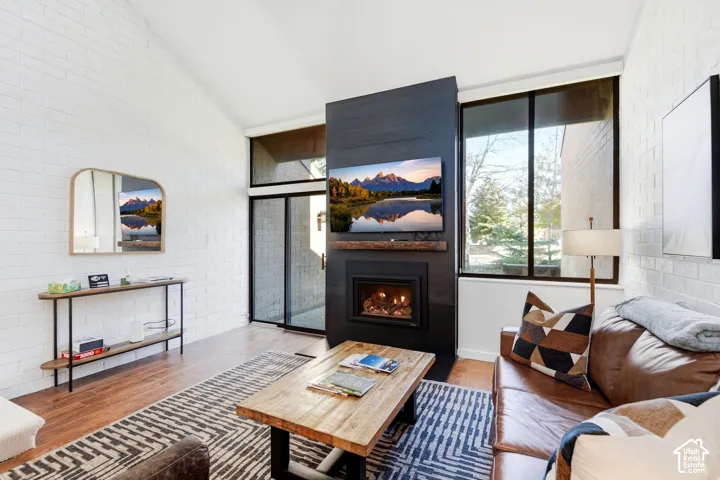 Vaulted ceiling, wood-finish flooring, white brick wall, modern fireplace, and large windows