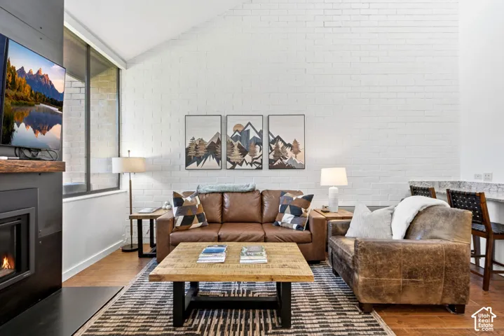 Vaulted ceiling, white painted brick wall, built-in fireplace with wood mantel, wood-finish flooring, and large window