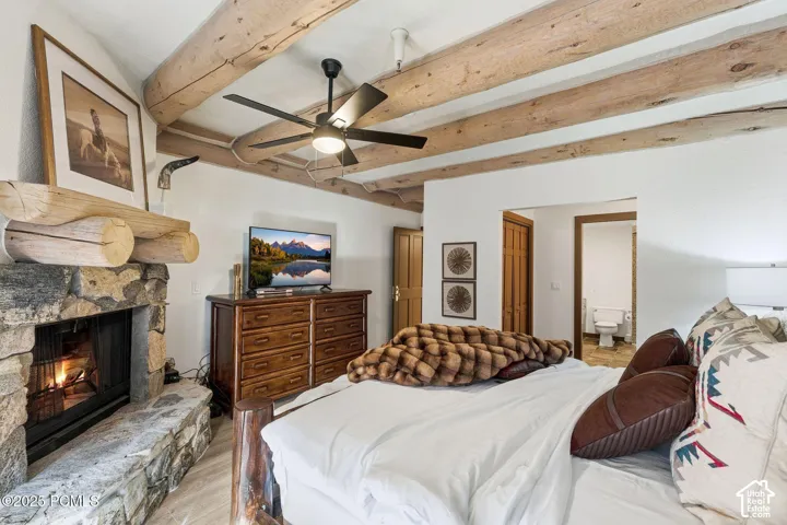 Primary Bedroom Suite with Exposed Beam Ceiling, Wood-Finish Dresser, Fireplace with Stone Surround, Ceiling Fan with Integrated Lighting, and Multiple Glass Doors Providing Natural Light Throughout