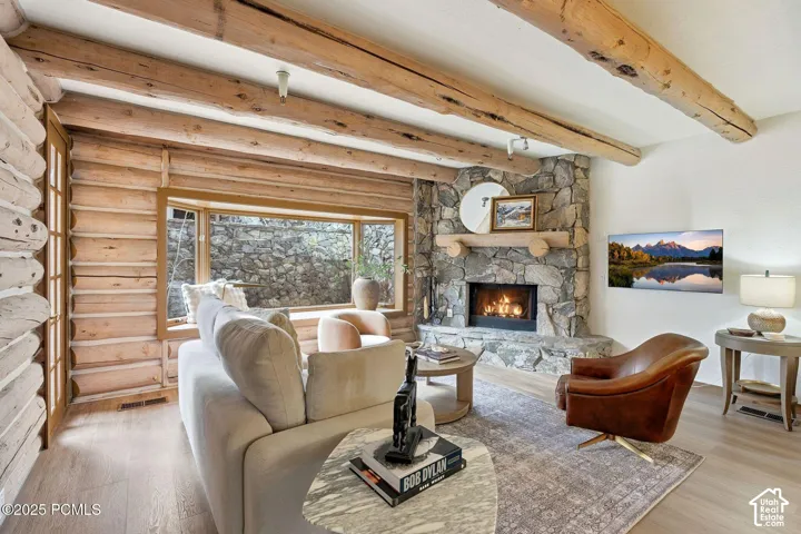 Exposed wood beam ceiling, stone-surround fireplace with a wood mantle, wood-finish flooring, and a bay window