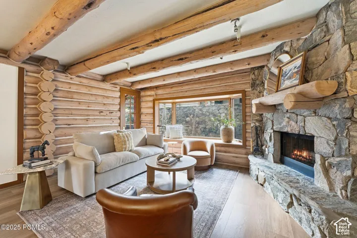 Rough-hewn log walls with exposed ceiling beams, stone fireplace with a log mantel, large window with a wood-framed sill, and wood-finish flooring