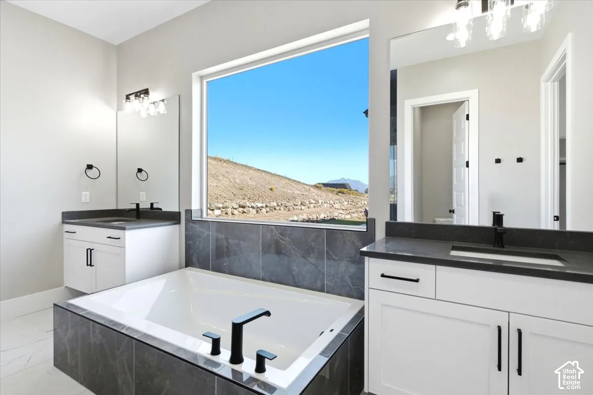 Freestanding soaking tub with dark tile surround, dual white vanities with dark countertops, and large framed window