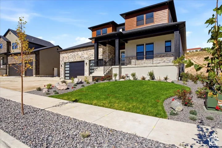 Modern exterior featuring brick and wood-tone siding, an attached garage with a contemporary door, a covered porch with a metal railing, and a landscaped front yard with a lawn and decorative rocks