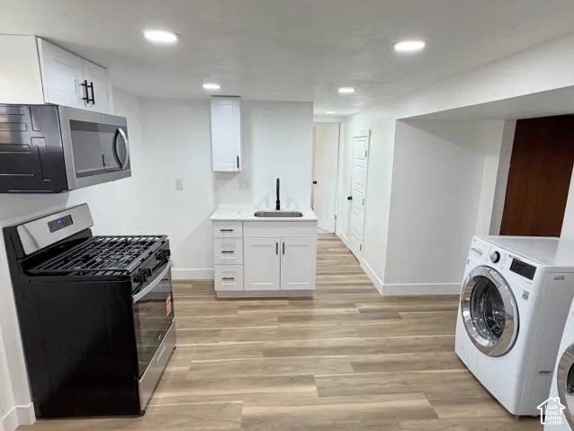 Contemporary kitchen and laundry area featuring wood-finish flooring, recessed lighting, white cabinetry with black hardware, stainless steel appliances, and a compact utility sink