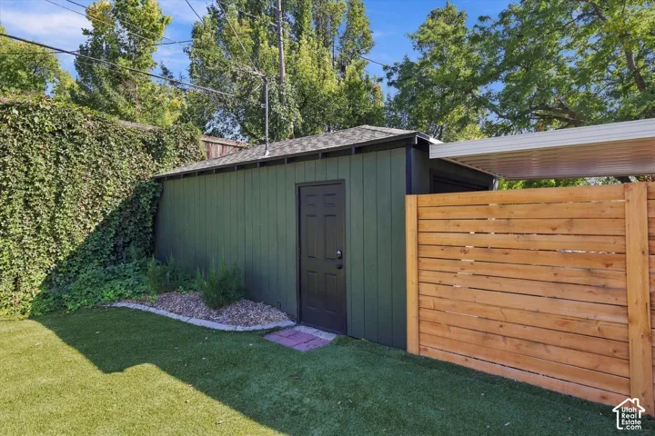 Detached structure with a dark shingle roof, natural wood privacy fencing, a covered carport extension, and a ground-level rock bed