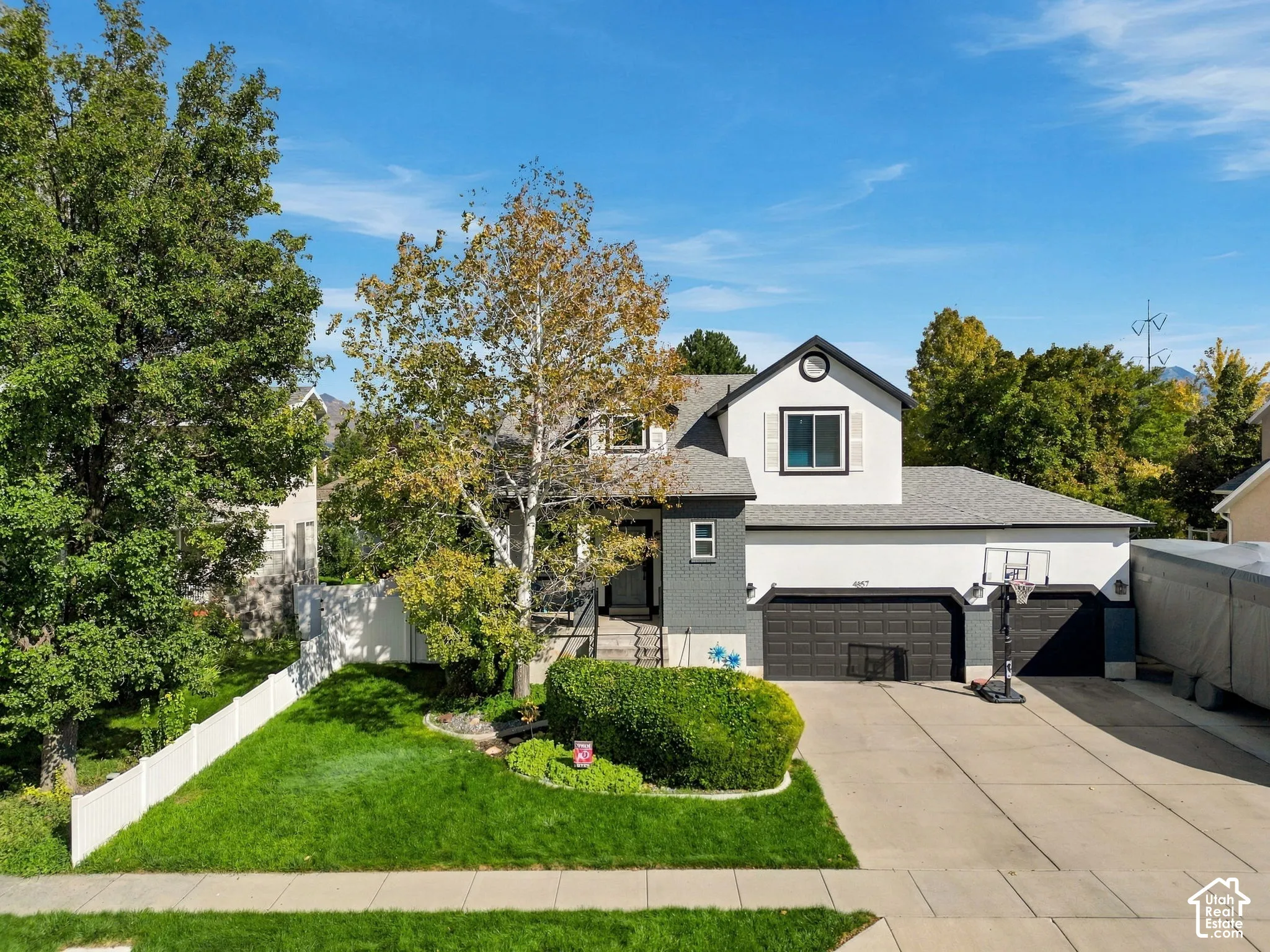 Two-story residence featuring a gray and white exterior, an attached two-car garage, mature landscaping, a paved driveway, and a white fence