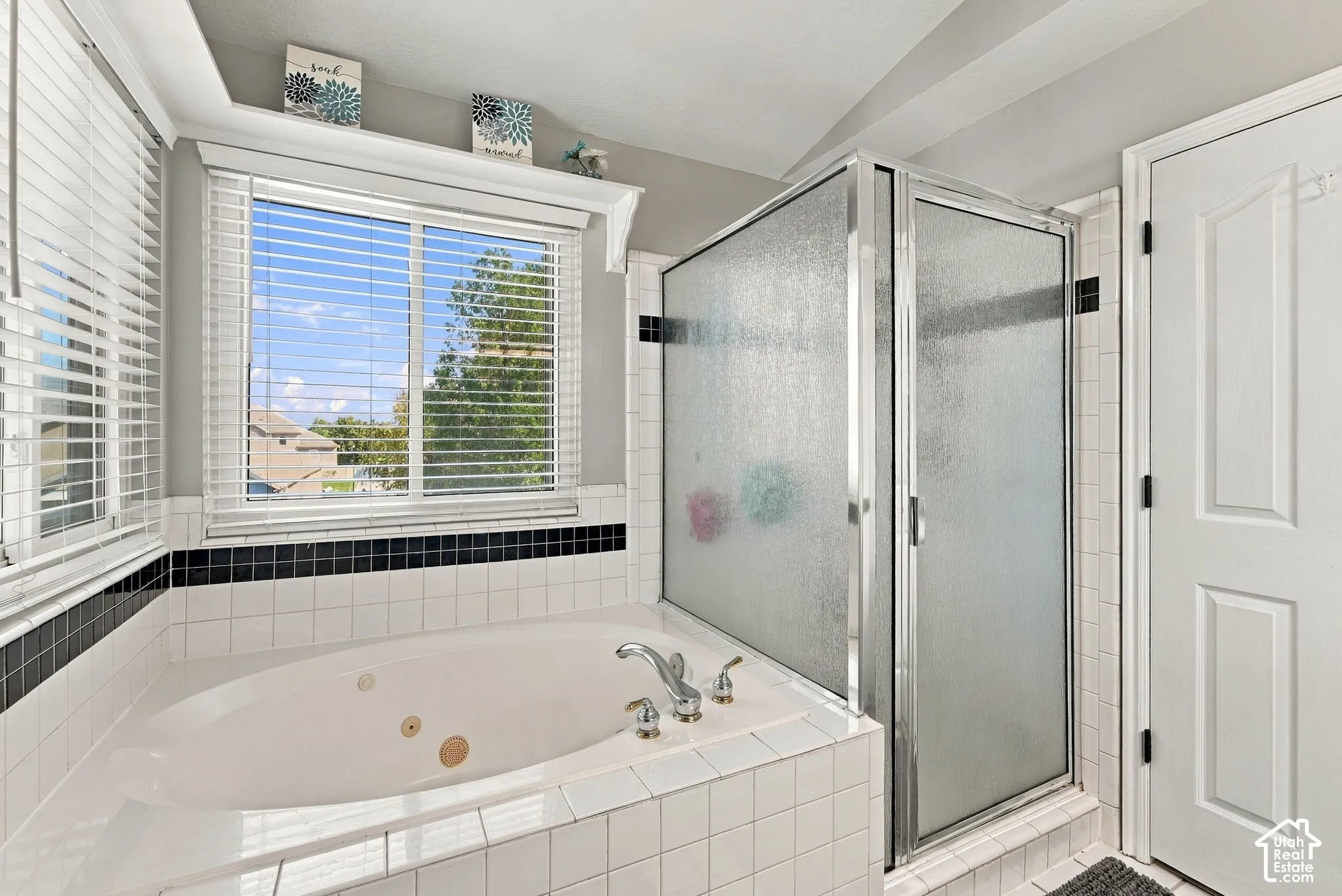 White tiled jetted tub with chrome fixtures, frosted glass shower enclosure, and windows with horizontal blinds