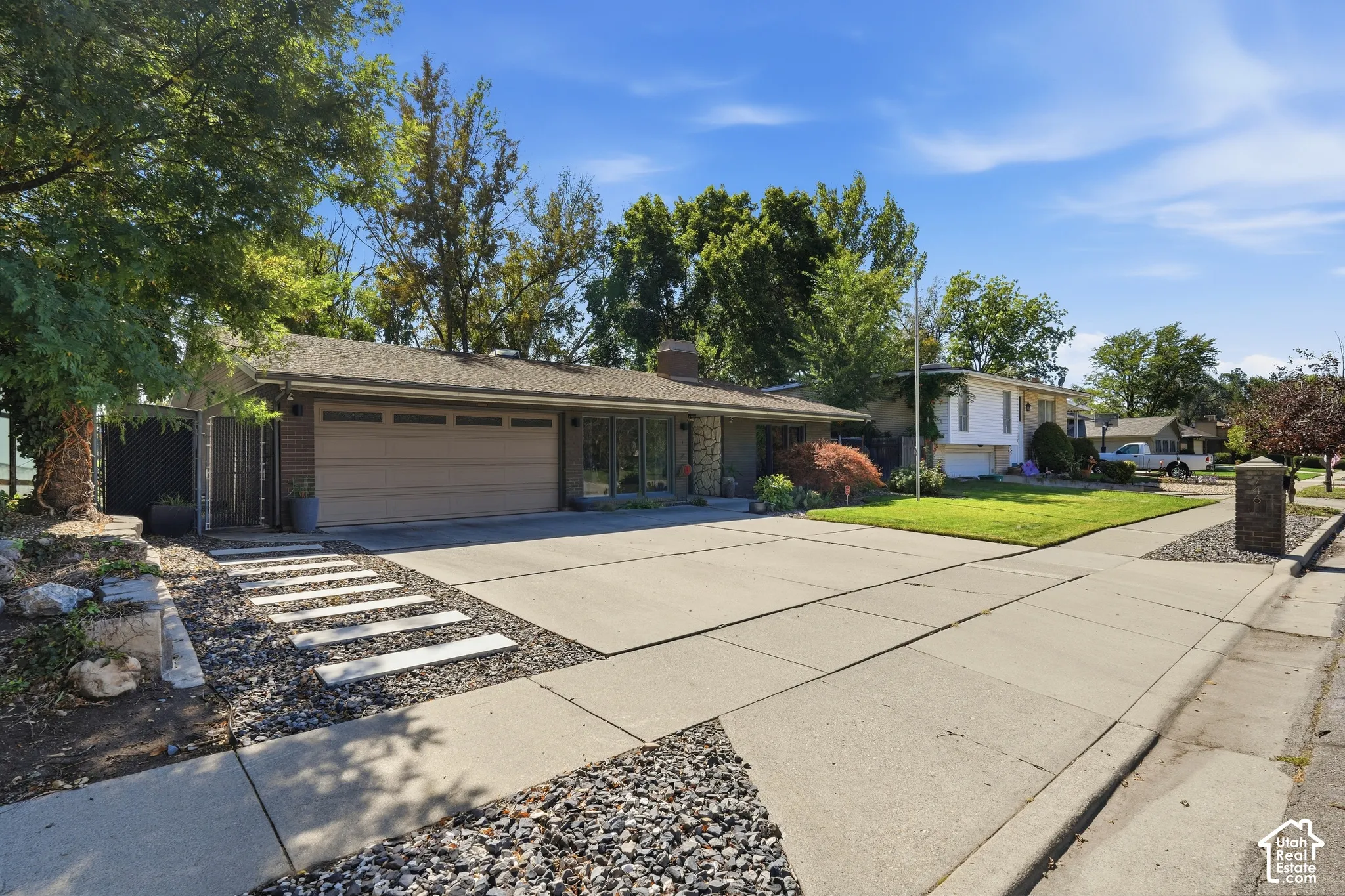 Expansive concrete driveway, attached garage, contemporary entry path with stepping stones, mature trees, and landscaped rock beds