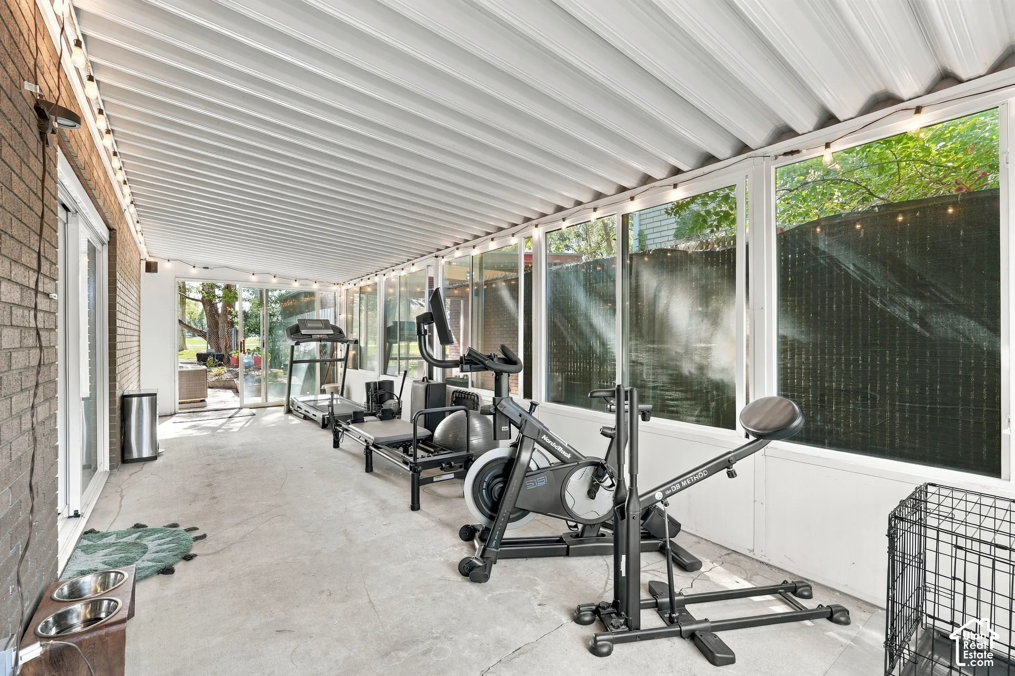 Covered outdoor fitness space featuring a corrugated metal roof, expansive concrete flooring, and large glass panels with black trim framing mature landscaping