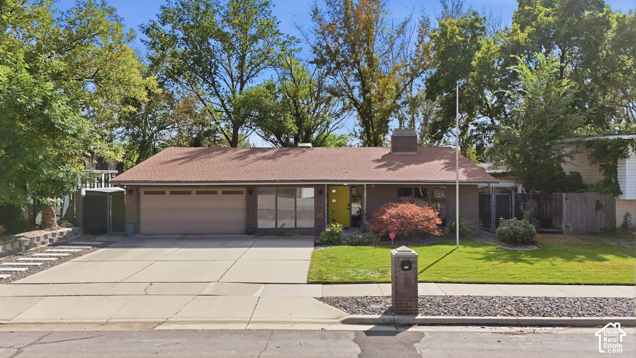 Single-story residence featuring a brown shingle roof, attached garage, paved driveway, and established landscaping with mature trees