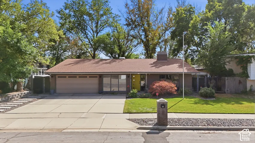 Single-story residence featuring a brown shingle roof, attached garage, paved driveway, and established landscaping with mature trees