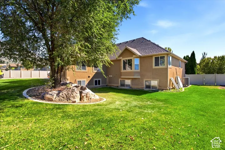 Expansive green lawn with a mature tree featuring a stone and mulch bed, stucco exterior, multiple window openings, and a perimeter fence