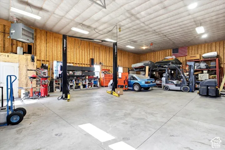 Spacious garage with concrete flooring, exposed wood-paneled walls, multiple overhead fluorescent light fixtures, and a vehicle lift system