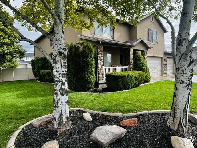 Two-story stucco exterior with stone accents, covered front porch with white railing, established shrubbery, and two-car garage