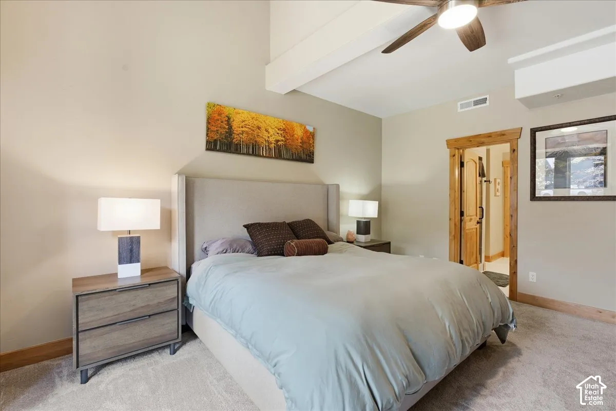 Vaulted ceiling with exposed beam, wood-finish ceiling fan, wood-trim doorway, upholstered headboard, and wood-finish nightstands