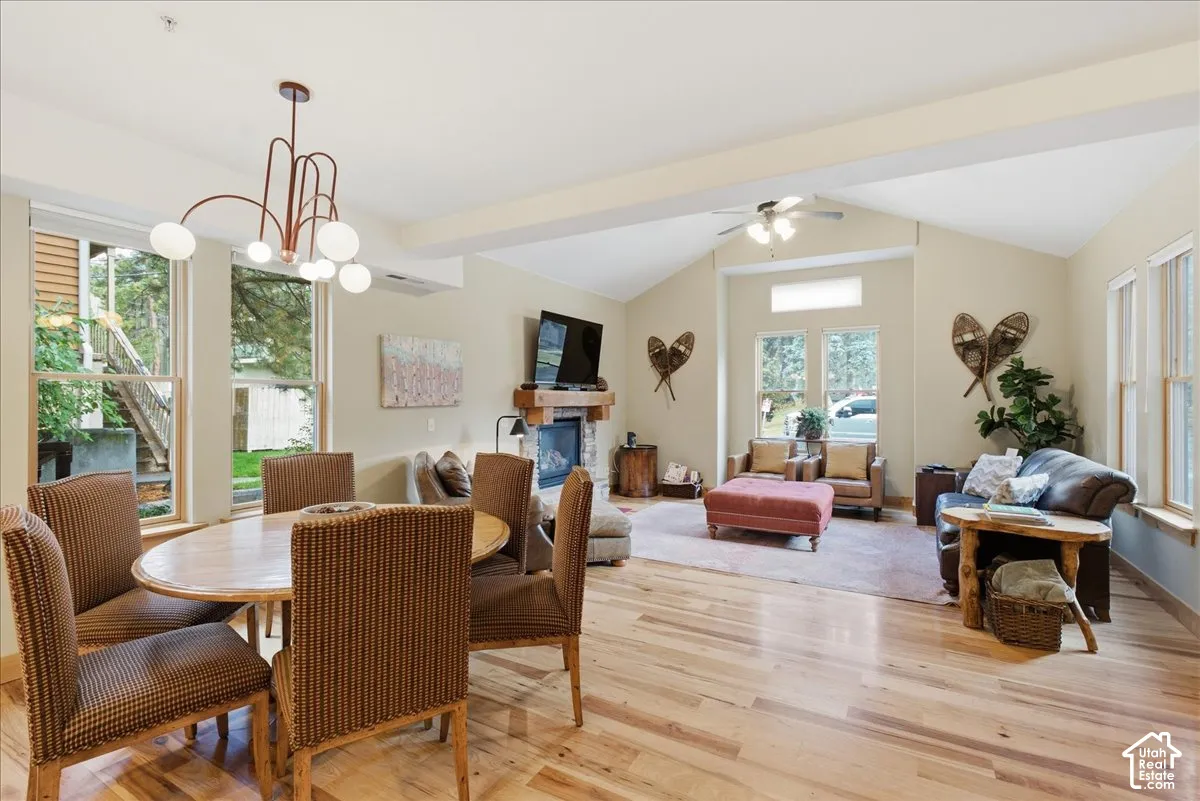 Vaulted ceiling with exposed beams, wood-finish flooring, stone-front fireplace with wood mantel, ceiling fan, and multiple large windows
