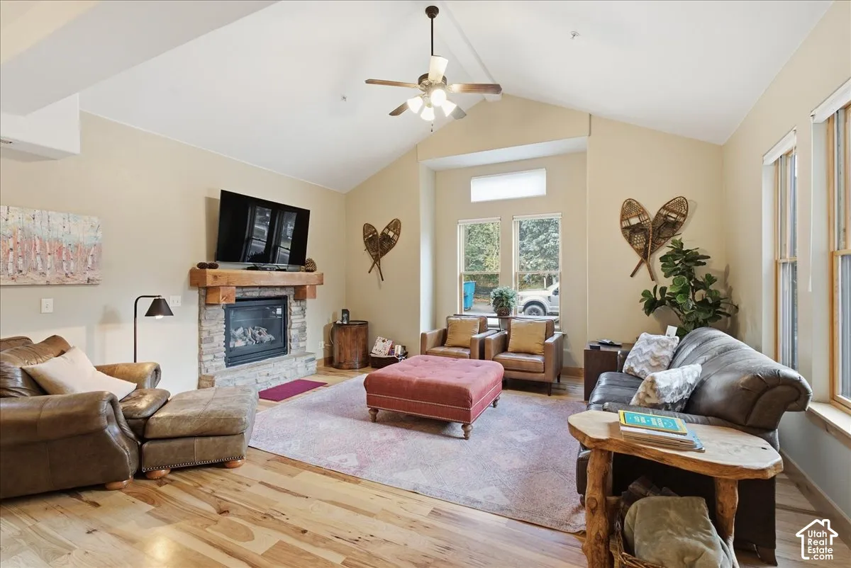Vaulted ceiling with a ceiling fan, stone fireplace with a wood mantle, large windows, and wood-finish flooring