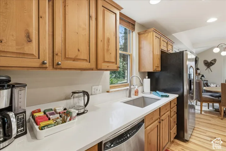 Wood-finish cabinetry, light-toned countertop, stainless steel sink with gooseneck faucet, stainless steel dishwasher, and wood-finish flooring