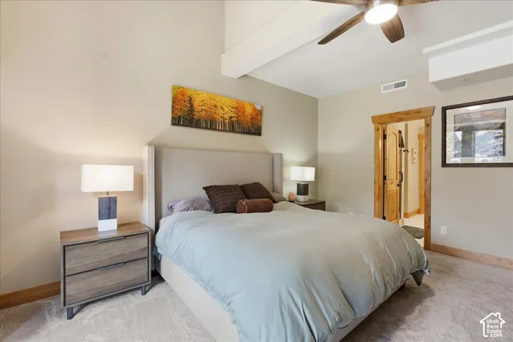 Vaulted ceiling with exposed beam, wood-finish ceiling fan, wood-trim doorway, upholstered headboard, and wood-finish nightstands