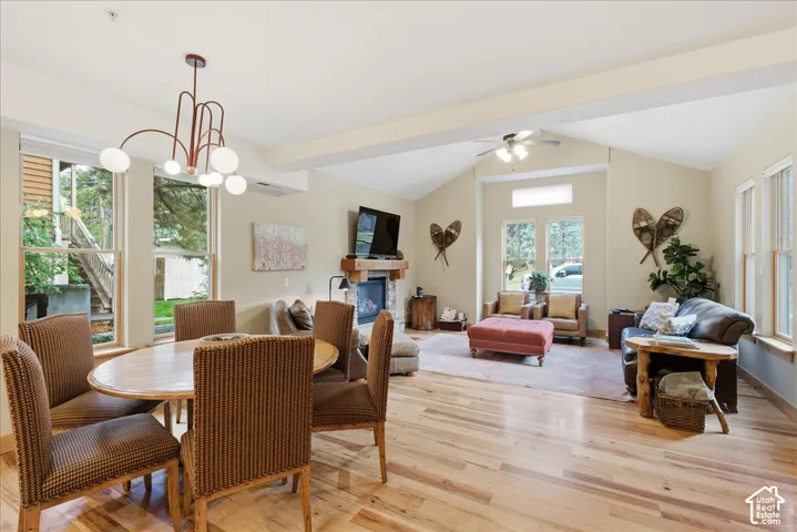 Vaulted ceiling with exposed beams, wood-finish flooring, stone-front fireplace with wood mantel, ceiling fan, and multiple large windows