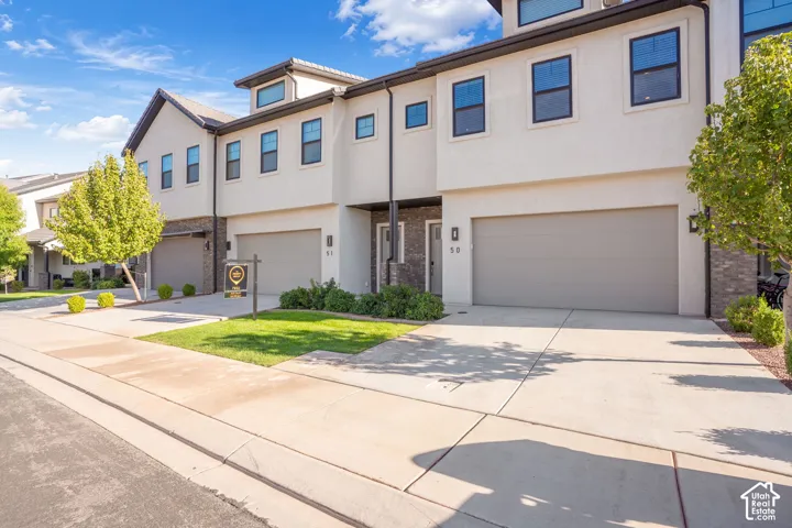 Modern townhome exterior with stucco and stone accents, featuring a two-car garage, concrete driveway, and neatly maintained landscaping